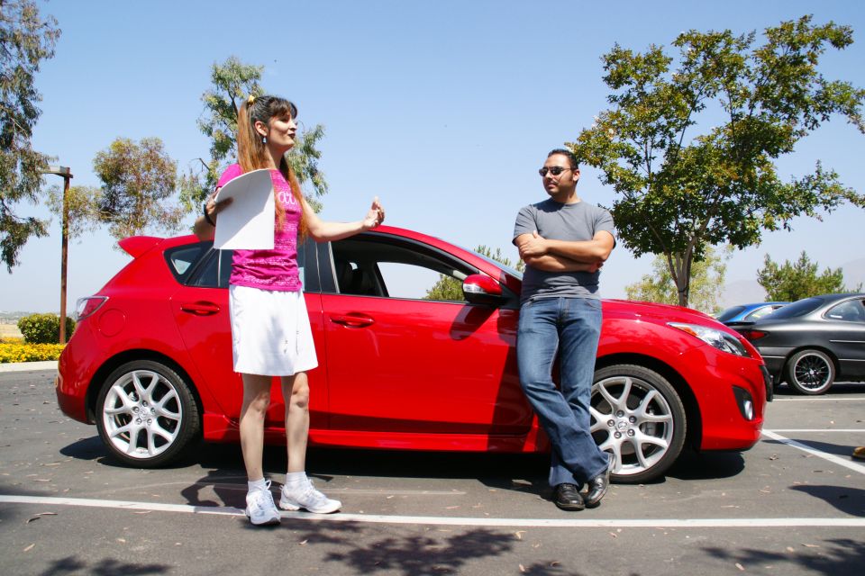 Woman in pink shirt and white skirt talks to man in gray shirt and jeans next to a red hatchback. Another car is parked behind