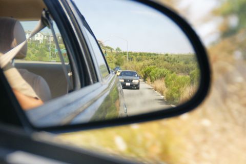 1995 Ford Taurus - A dark sedan is visible in the side mirror of another vehicle, driving on a road with dry brush and trees.