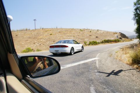 1995 Ford Taurus - A white Ford Probe is seen from behind driving on a winding road through a dry, hilly landscape.