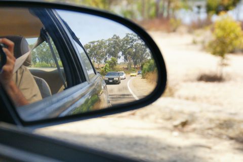 1995 Ford Taurus - A view through a car's side mirror shows multiple vehicles traveling on a road surrounded by dry brush and trees.