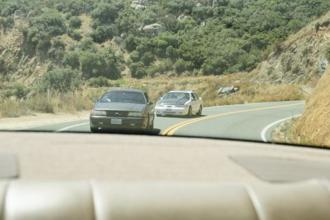 1995 Ford Taurus - Two cars, a gray 1995 Ford Taurus and a white car, drive on a winding road through a hilly, arid landscape.