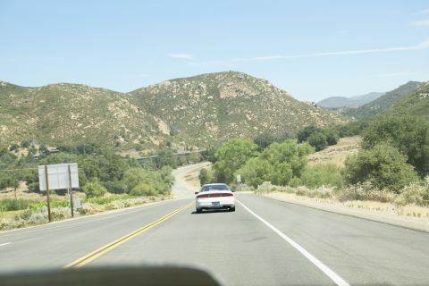 1995 Ford Taurus - A white 1995 Ford Taurus travels down a highway, surrounded by rolling hills and trees under a clear blue sky.