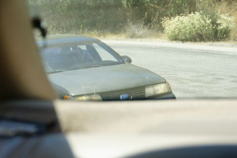 1995 Ford Taurus - A faded sedan with a blue emblem sits on the shoulder of a road, surrounded by dry brush.