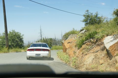 1995 Ford Taurus - A white sedan drives down a winding road bordered by rocky terrain and sparse vegetation under a clear blue sky.