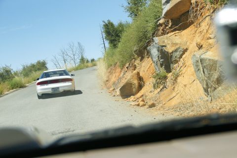 1995 Ford Taurus - A white 1995 Ford Taurus is seen from behind as it drives up a winding road bordered by rocky terrain and vegetation.