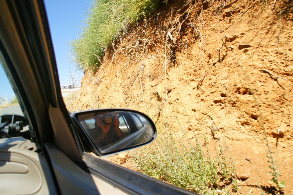 View from inside a car, looking out the passenger window at a dirt embankment and vegetation. Side mirror reflects the driver holding a camera