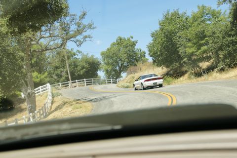 1995 Ford Taurus - A white 1995 Ford Taurus sedan navigates a curve on a rural road with trees and a white fence.