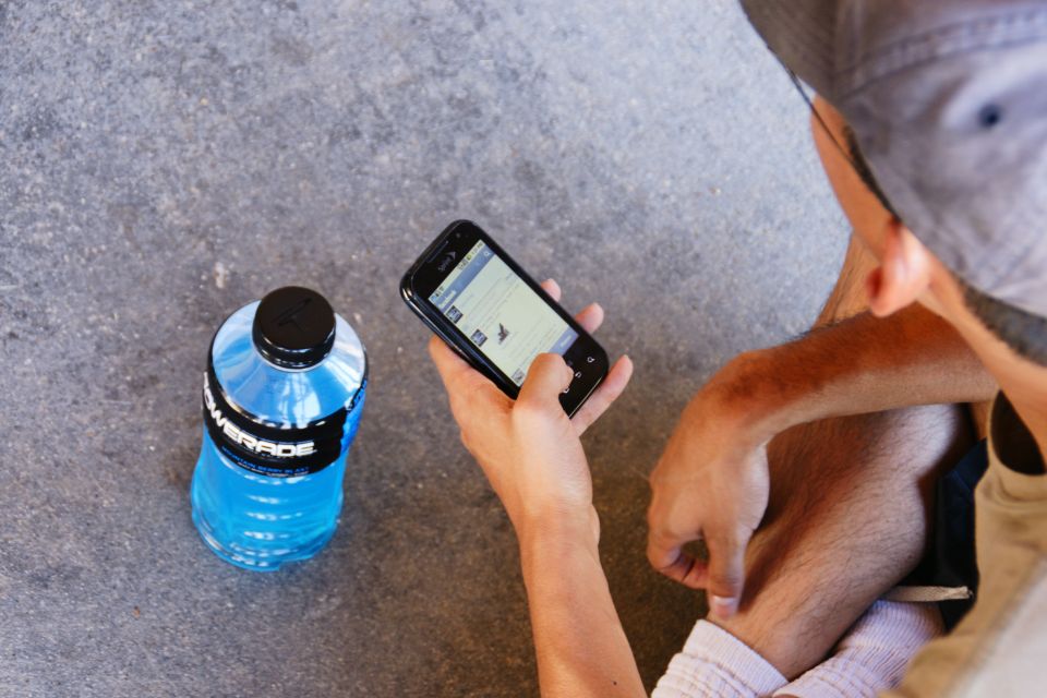 Person holding smartphone with social media feed, next to blue Powerade bottle on concrete