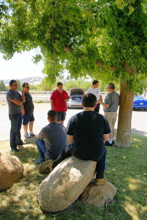 1995 Ford Taurus - A group of men are gathered around a silver sedan with its hood open, parked outdoors on a sunny day.