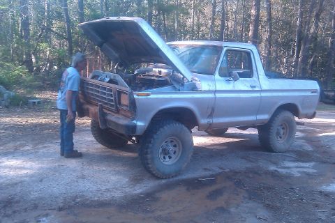 1978 Ford Bronco - A man looks at the engine of a silver 1978 Ford Bronco parked on a dirt road.