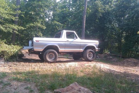1978 Ford Bronco - A silver 1978 Ford Bronco sits off-road, surrounded by dense foliage and trees.