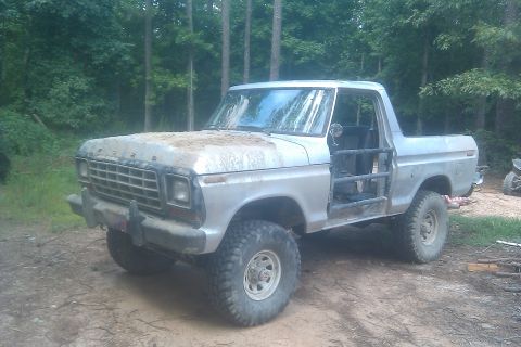 1978 Ford Bronco - A modified 1978 Ford Bronco sits on a dirt path surrounded by trees, showing its roll cage and large tires.