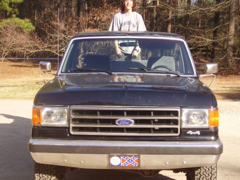 1988 Ford Bronco - A person stands atop a black Ford Bronco, which is parked outdoors with trees in the background.