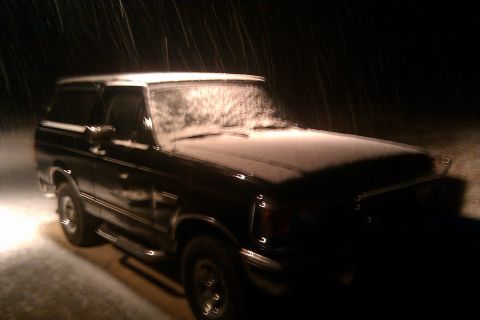 1988 Ford Bronco - A dark-colored Ford Bronco is visible in the snow at night, with its front end illuminated by a light source.
