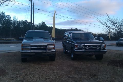 1988 Ford Bronco - Two SUVs, a Ford Bronco and a Chevrolet pickup, are parked on a lot under a cloudy sky.