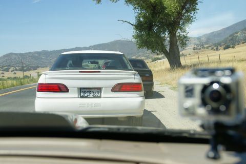 1995 Ford Taurus - A white 1995 Ford Taurus is followed by another vehicle on a rural road under a clear blue sky.