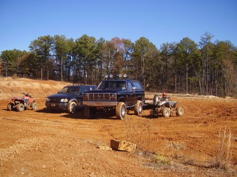 1986 Ford Bronco - Two SUVs and ATVs are parked on a dirt lot with trees in the background under a clear blue sky.