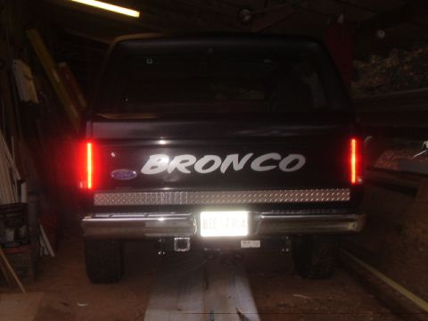 1986 Ford Bronco - The rear of a black 1986 Ford Bronco is visible, vertical taillights and a diamond plate bumper accent.