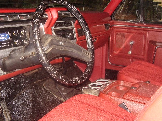 Interior of a 1986 Ford Bronco with red seats and dashboard, showing the steering wheel and center console