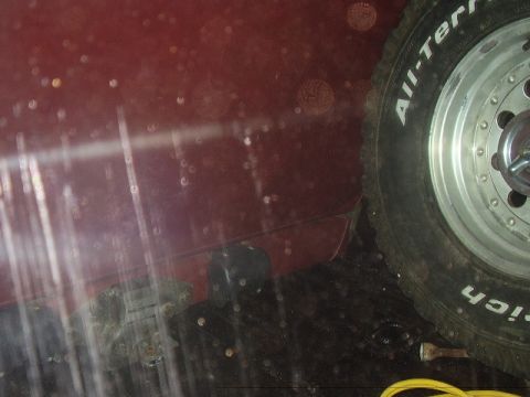 1986 Ford Bronco - The side of a red vehicle and its tire are visible, with water droplets on the surface.