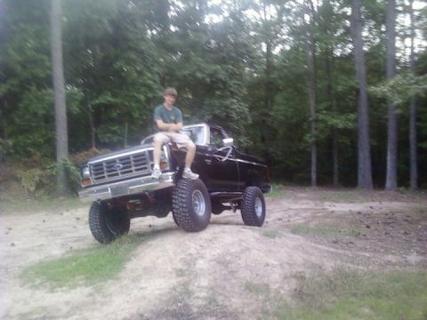 1986 Ford Bronco - A person is perched on the front of a modified pickup truck, which is positioned on a small hill outdoors.