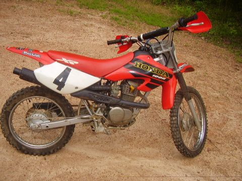 1986 Ford Bronco - Side view of a red Honda 100R dirt bike parked on a dirt surface.