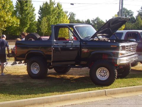 1986 Ford Bronco - This lifted Ford Bronco is parked on grass with its hood up, showing the engine bay.