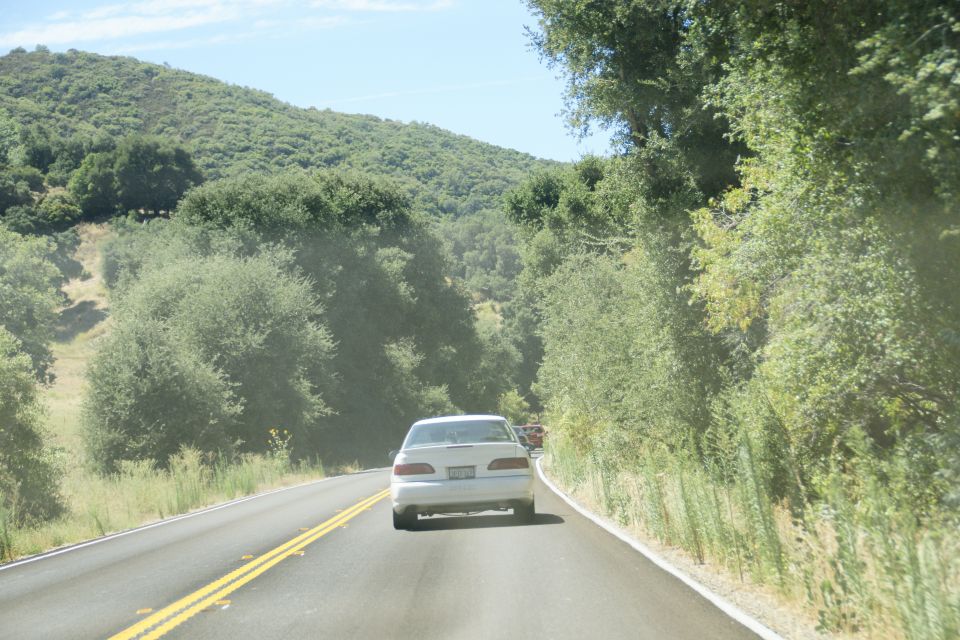 White sedan driving on a two-lane road through a hilly, tree-covered area. The car is seen from behind