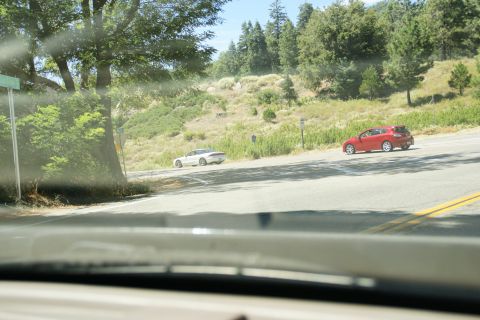 1995 Ford Taurus - Two cars, a white sports car and a red hatchback, drive along a scenic, winding road bordered by trees and hills.