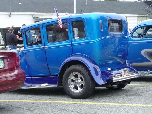 Bright blue prewar Ford sedan street rod, rear three-quarter, on chrome wheels in a paved car-show lot
