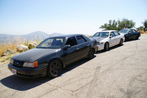 1995 Ford Taurus - Three sedans are parked on a scenic mountain road with dry grass and hills in the background.