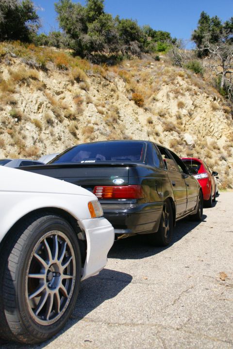 1995 Ford Taurus - Three cars are parked on a paved road next to a dry, rocky hillside under a clear blue sky.