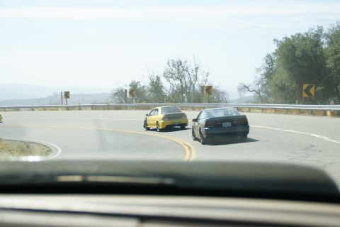 1995 Ford Taurus - Two cars navigate a sharp curve on a scenic mountain road, with yellow and black vehicles visible.