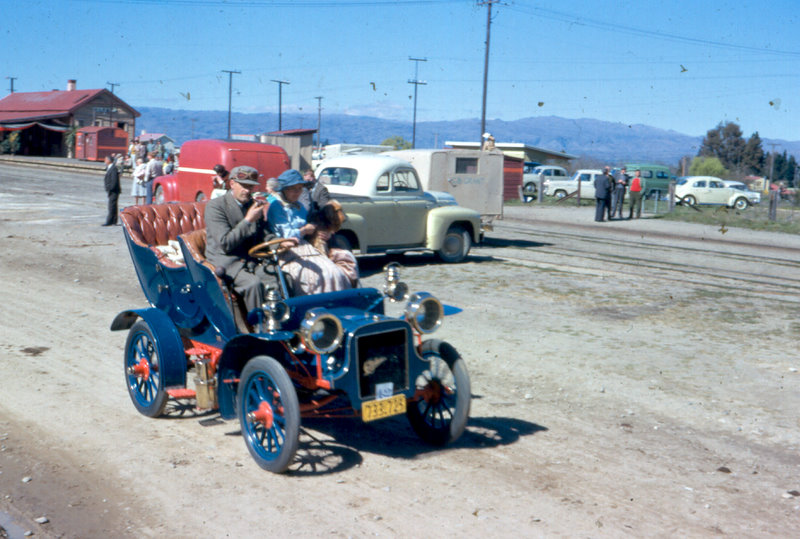 Blue brass-era Edwardian tourer with red wheels driven on a dirt road, vintage colour photo