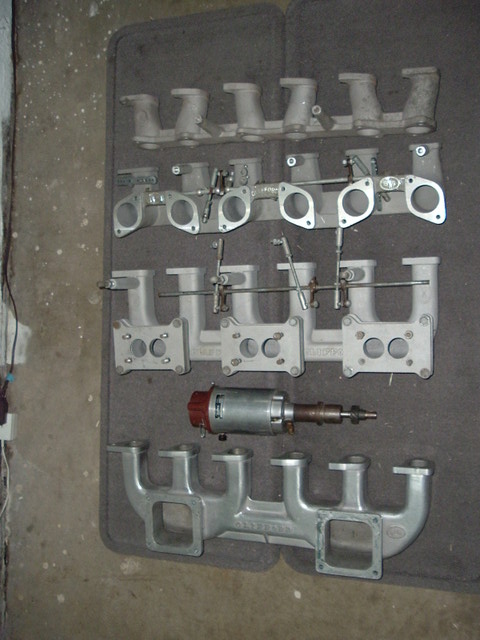 1981 Ford Bronco - Several cast aluminum intake manifolds and carburetor adapter plates with linkage rods laid out in rows on a gray moving blanket on a concr…