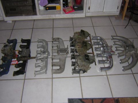 1981 Ford Bronco - Six inline six-cylinder manifolds arranged side by side on a white tile floor, ranging from blue and cast-iron pieces on the left to a trip…