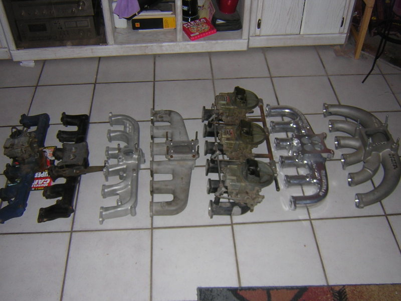 Six inline-six intake and header manifolds arranged on tile floor, one with triple sidedraft carbs