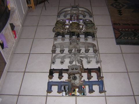 1981 Ford Bronco - A row of inline six-cylinder intake and exhaust manifolds lined up on a tile floor for comparison, including a triple-carb aluminum setup,…