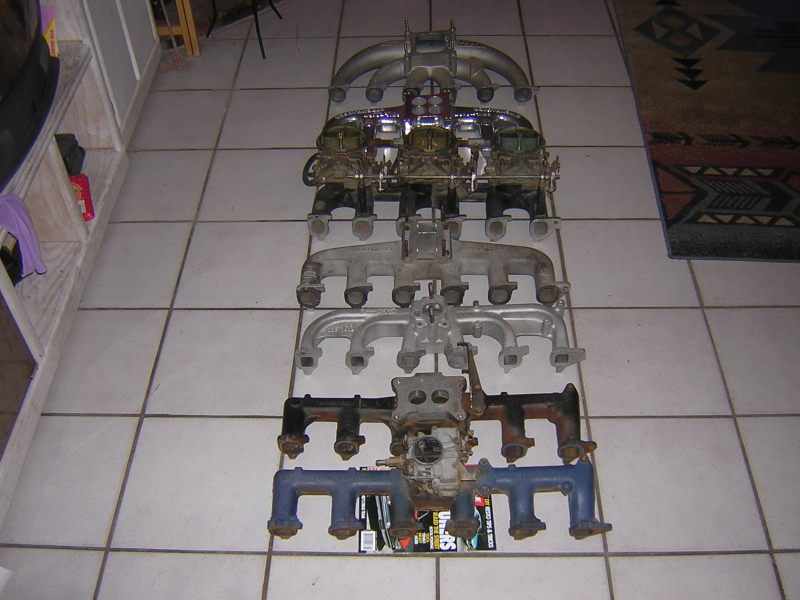 Row of inline-six intake and exhaust manifolds on tile floor, including a triple-carb setup