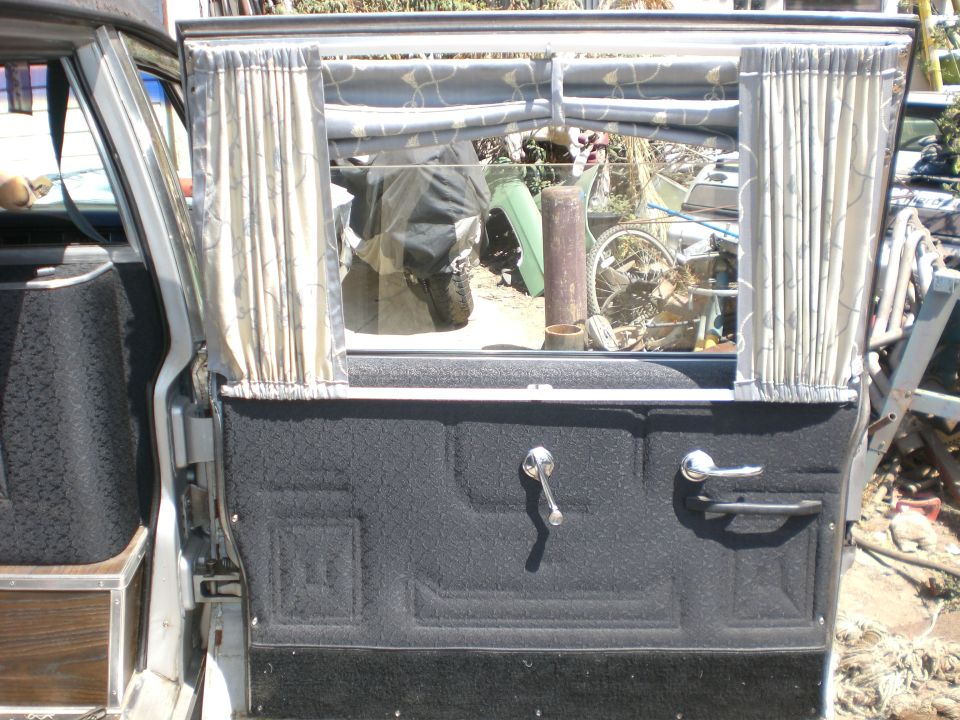 Open rear door interior with black upholstered panel, chrome handles, and cream curtains on a Cadillac Fleetwood