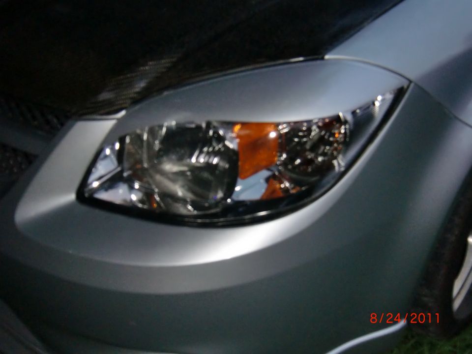 Car's front headlight and bumper area. Date and website visible