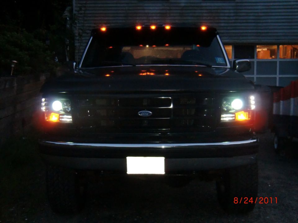 Front of a dark Ford F-Series truck with headlights and amber marker lights on. It has a chrome bumper and a blank license plate