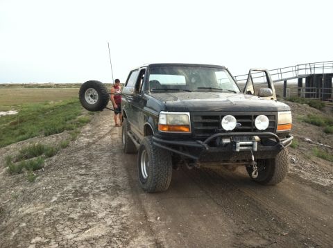 1994 Ford Bronco - A black Ford Bronco is parked on a dirt path with a spare tire mounted on the rear carrier.