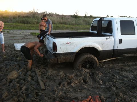1994 Ford Bronco - A white pickup truck is stuck deep in mud, with several people attempting to push it free.