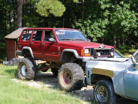 1978 Ford Bronco - A modified red Jeep Cherokee sits high on oversized tires, parked beside a light blue pickup truck in a grassy area.