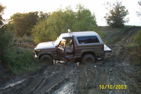 1982 Ford Bronco - A brown and white Ford Bronco is stuck in deep mud on an off-road trail, with its doors open.