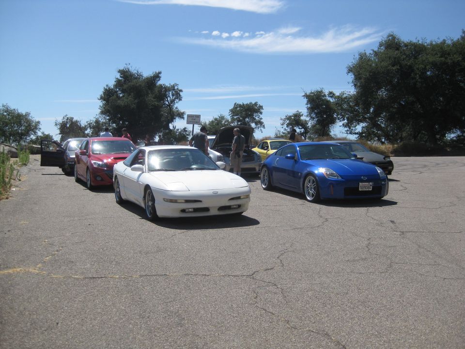 Several cars parked on asphalt under a blue sky, including a white coupe and a blue sports car