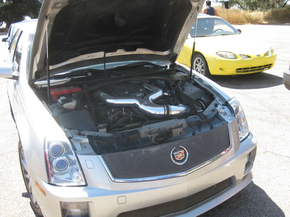 Silver Cadillac CTS-V with hood open showing engine and intake piping. Yellow car in background
