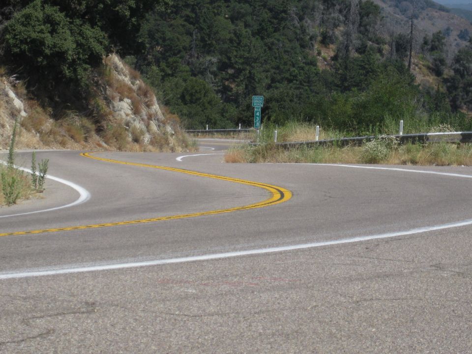 Winding mountain road with double yellow lines and guardrail. Signpost visible