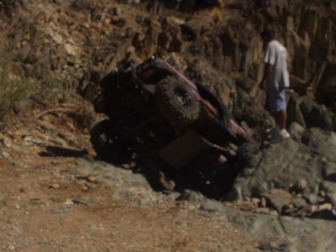1993 Ford Bronco - An off-road vehicle has tipped onto its side on a rocky incline, with a person observing the scene.